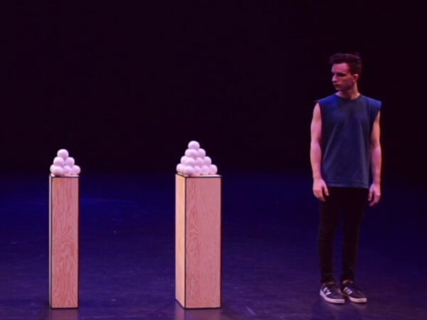 Juggler Domenyk La Terra standing next to two pyramids of juggling balls, each pyramid atop a pillar.