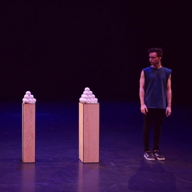 Juggler Domenyk La Terra standing next to two pyramids of juggling balls, each pyramid atop a pillar.